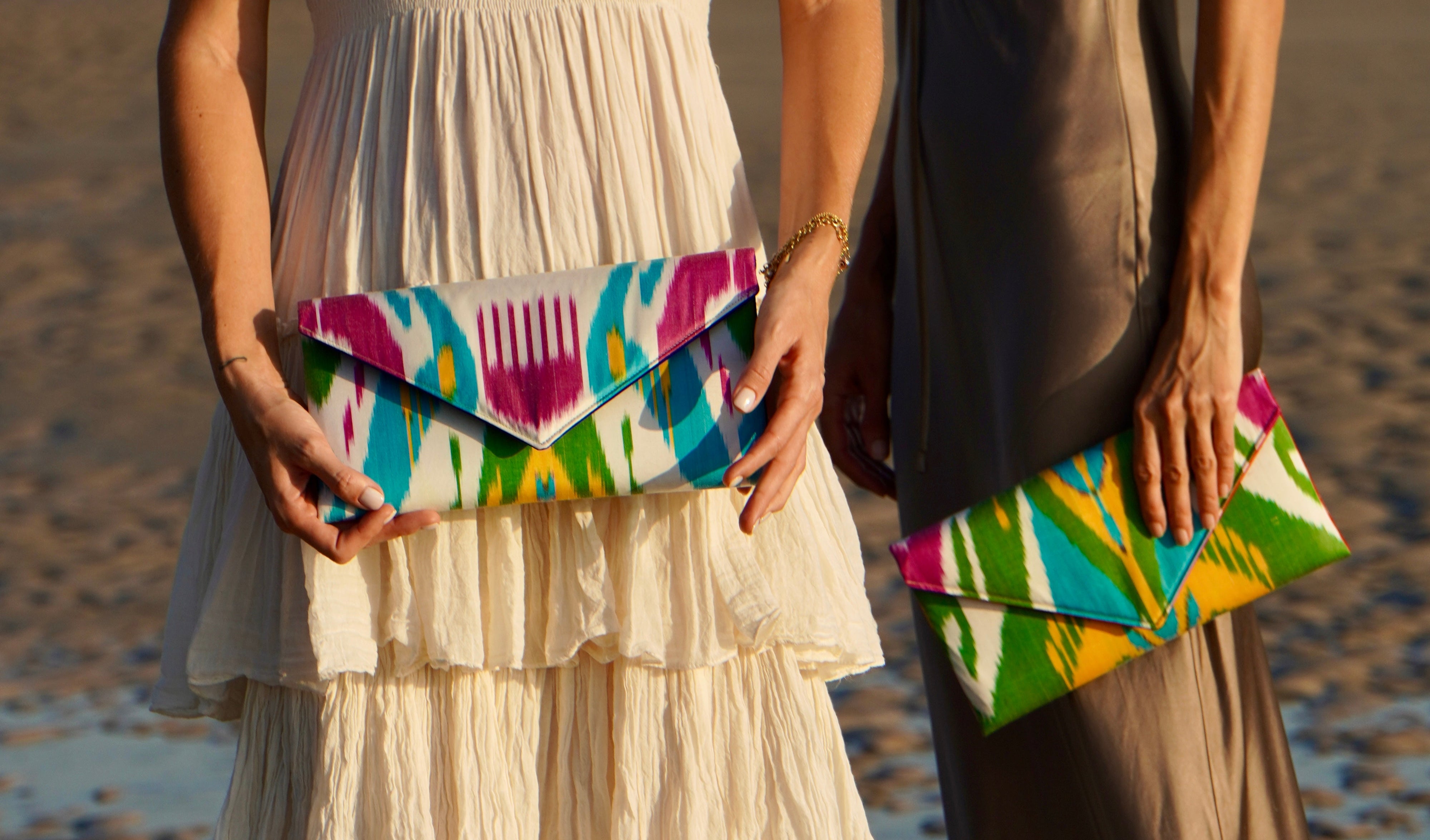 Two women on a beach holding colorful Frankitas silk ikat handwoven clutch bags.