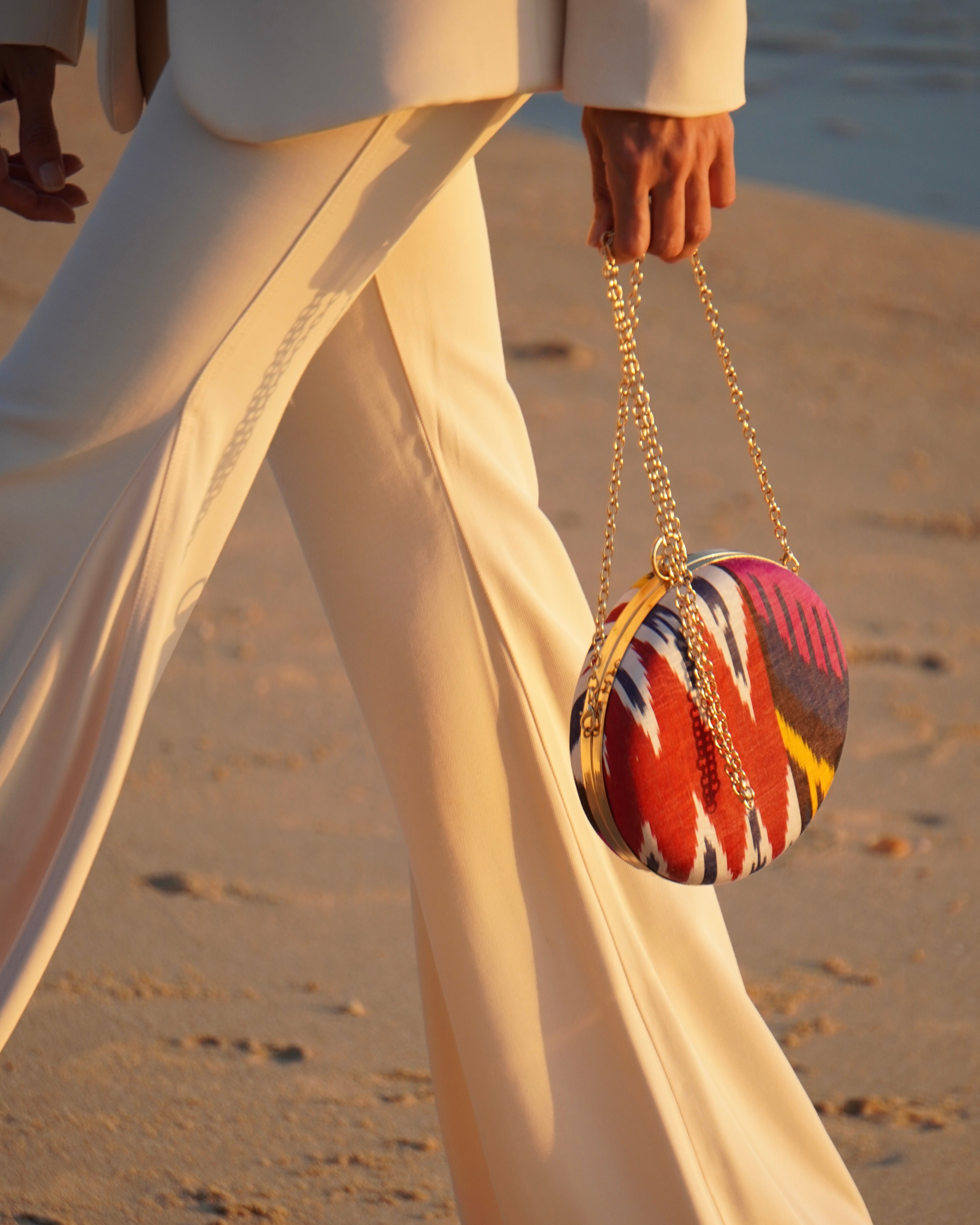 Woman holding a colorful round metal silk ikat handwoven clutch handbag on a sandy beach