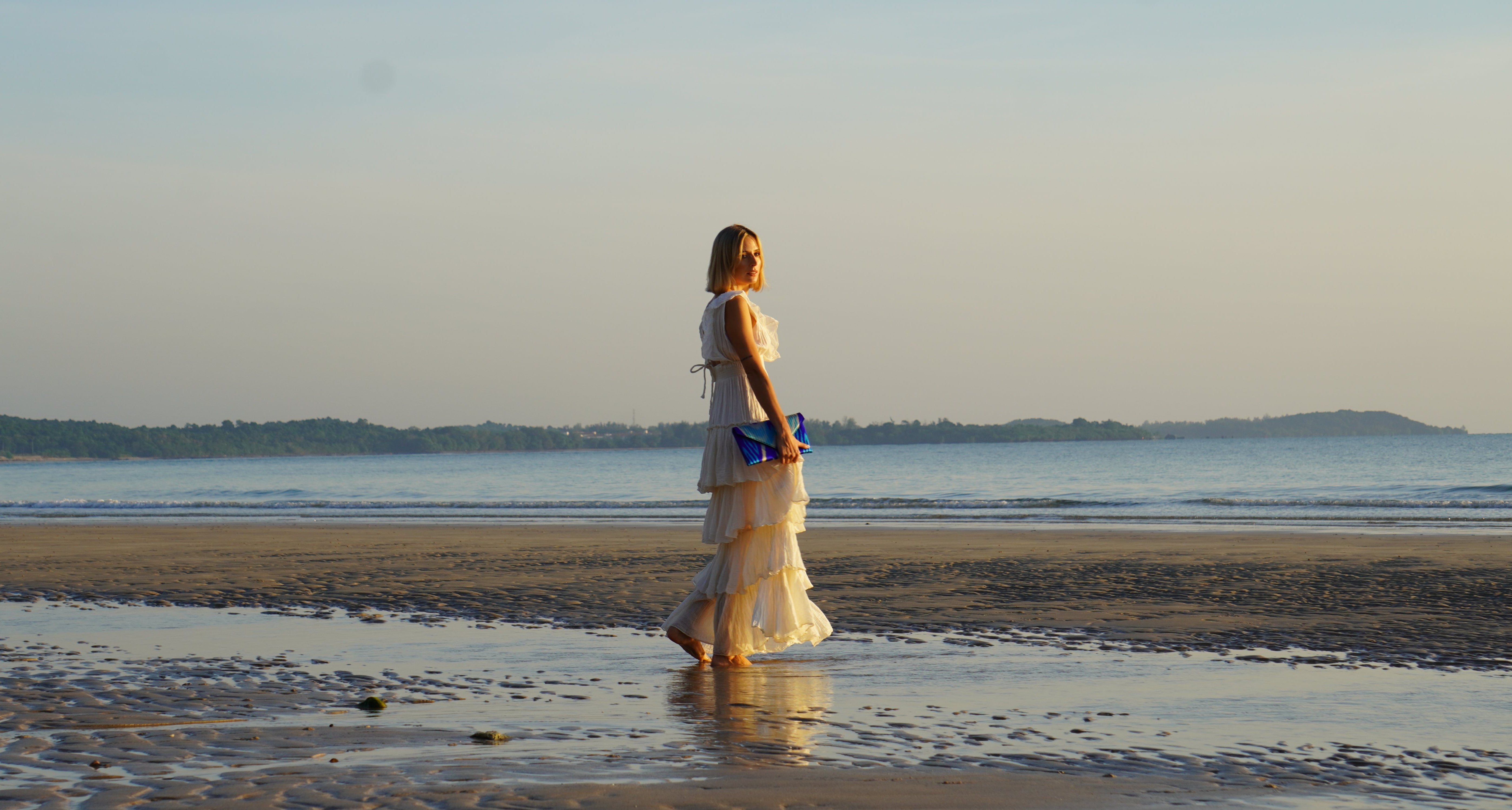 Woman in a white dress standing on a beach holding a  Frankitas blue silk ikat handwoven clutch handbag